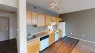 This single-floor residential unit offers a practical and functional layout designed for comfortable everyday living. The main living area is bright and open, featuring neutral-toned carpet, light beige walls, and a textured ceiling. Large windows allow abundant natural light to fill the space while framing views of the surrounding urban cityscape, creating an airy and connected atmosphere.

The kitchen is laid out in a classic galley style, finished with warm honey-toned wood cabinetry that extends to the upper walls for generous storage capacity. Countertops provide ample prep space, and the kitchen is equipped with a full suite of white appliances including a coil electric range, dishwasher, and refrigerator. Track lighting overhead illuminates the workspace effectively. Rich dark hardwood-style flooring in the kitchen and adjacent areas provides a grounded contrast to the carpeted living space.

The bathroom is clean and efficiently designed, featuring a full tub and shower combination with a chrome curtain rod, a single-sink vanity with wood cabinetry, and hardwood-style flooring that ties back to the rest of the unit. Overall, the space maintains a cohesive, neutral palette throughout, offering a move-in-ready environment suited to a range of lifestyles.