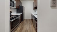 Located in the heart of Springfield, MA, this unit at Silverbrick Lofts combines industrial-chic character with modern residential comfort. The open living and dining areas are defined by rich, dark hardwood-style flooring, crisp white walls, and distinctive black track lighting that runs along the ceiling — a hallmark of the loft aesthetic. Exposed structural elements add an urban, warehouse-inspired character throughout the space. The galley-style kitchen is well-appointed with dark espresso cabinetry, white countertops, and a full suite of stainless steel appliances, including a refrigerator, range, microwave, and dishwasher, providing a sleek and functional cooking environment. The bedroom features lighter hardwood-style flooring, track lighting, and an open closet with built-in rod, offering practical storage. The bathroom is finished with large-format neutral tile surrounding a full soaking tub and shower, complemented by a dark wood vanity with a white basin. Throughout the unit, the clean lines, neutral palette, and contemporary fixtures create a cohesive, modern ambiance that reflects the loft's thoughtful design sensibility.