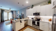 This thoughtfully designed single-floor residence combines modern aesthetics with functional living. The open-concept kitchen serves as the heart of the home, showcasing sleek shaker-style cabinetry in slate gray and blue-gray tones, granite countertops, stainless steel appliances, and a generously sized island with bar seating. Pendant lighting adds a contemporary touch, while wood-look flooring flows seamlessly throughout the space. The kitchen opens directly to the living area, which features large windows that invite natural light and views of the outdoors, creating an airy, connected atmosphere.

The primary bedroom offers a sophisticated retreat with a bold navy accent wall, large windows with draping curtains, and plush carpeting. A second bedroom maintains a softer, neutral palette with ample natural light. Both bathrooms are well-equipped with granite-topped vanities, gray cabinetry, wood-look flooring, and direct access to dedicated walk-in closets, providing excellent storage and convenience.

The living area rounds out the space with generous wall space ideal for artwork display, complementing the overall contemporary design. Throughout, the residence maintains a cohesive, upscale character defined by quality finishes, thoughtful layout, and a harmonious blend of bold accent colors against clean, neutral backgrounds.