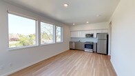 This single-floor space showcases a clean, contemporary design with an open-concept layout that seamlessly connects the living area to the kitchen. The interior is finished with light hardwood flooring throughout, contributing to a warm yet modern aesthetic. White walls and recessed lighting enhance the bright, airy atmosphere of the space.

The kitchen is well-appointed with a full suite of stainless steel appliances, including a refrigerator, range with oven, built-in microwave, and dishwasher. Cabinetry features a two-tone design — crisp white upper cabinets paired with warm wood-toned lower cabinets — providing ample storage while adding visual depth. A sink with a tiled backsplash completes the functional kitchen setup.

The living area is generously proportioned, offering an open and flexible space ideal for a variety of furniture arrangements. A large bank of windows along one wall floods the room with natural light and frames views of mature trees and surrounding greenery, creating a pleasant indoor-outdoor connection. The clean lines, neutral palette, and quality finishes throughout give this space a fresh, move-in-ready feel that appeals to a wide range of occupants.