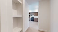 This thoughtfully designed single-floor unit offers a clean, modern aesthetic with consistent light wood-tone flooring running throughout the main living areas. The contemporary kitchen is a standout feature, outfitted with warm wood-grain cabinetry that extends to the ceiling for generous storage, a black electric range, a stainless steel refrigerator, and light-colored countertops. The kitchen opens naturally toward the main living area, creating an efficient and connected layout. Built-in open shelving near the entry provides additional practical storage and adds an architectural detail to the space. A clean, well-lit hallway connects the main areas, leading to both the bathroom and bedroom. The bathroom is finished with wood-toned cabinetry, a single vanity with a mirrored cabinet, a tub and shower combination with a curtain rod, and the same light wood flooring carried throughout the unit. The bedroom is bright and carpeted, featuring a window with blinds that allows natural light to pour in, casting warm light across the room. Crisp white walls and simple, modern trim work are consistent throughout, lending the unit a fresh, move-in-ready feel suited to a variety of lifestyles.