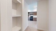 This thoughtfully designed single-floor unit offers a clean, modern aesthetic with consistent light wood-tone flooring running throughout the main living areas. The contemporary kitchen is a standout feature, outfitted with warm wood-grain cabinetry that extends to the ceiling for generous storage, a black electric range, a stainless steel refrigerator, and light-colored countertops. The kitchen opens naturally toward the main living area, creating an efficient and connected layout. Built-in open shelving near the entry provides additional practical storage and adds an architectural detail to the space. A clean, well-lit hallway connects the main areas, leading to both the bathroom and bedroom. The bathroom is finished with wood-toned cabinetry, a single vanity with a mirrored cabinet, a tub and shower combination with a curtain rod, and the same light wood flooring carried throughout the unit. The bedroom is bright and carpeted, featuring a window with blinds that allows natural light to pour in, casting warm light across the room. Crisp white walls and simple, modern trim work are consistent throughout, lending the unit a fresh, move-in-ready feel suited to a variety of lifestyles.