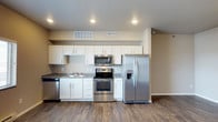 This single-story space showcases a well-appointed kitchen as its focal point. The kitchen is outfitted with a full suite of stainless steel appliances, including a French door refrigerator with an in-door water and ice dispenser, an electric range with oven, an over-the-range microwave, and a dishwasher. White shaker-style cabinetry extends from counter height to the ceiling, maximizing storage capacity, while granite-look countertops provide functional and attractive work surfaces. A double-basin sink is positioned beneath upper cabinets along the side wall. The flooring throughout is a durable wood-style laminate in a warm, medium-toned finish, adding visual warmth to the space. Recessed lighting in the ceiling provides even illumination across the kitchen area. Neutral beige wall tones complement the white cabinetry and create a clean, cohesive aesthetic. HVAC vents are integrated into the ceiling and upper wall, ensuring efficient climate control. The open layout between the kitchen and adjacent living area contributes to a spacious, connected feel throughout the unit.