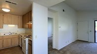 This single-floor residential space offers a functional and practical layout designed for comfortable everyday living. The kitchen is positioned at the front of the unit and features warm wood-toned cabinetry, a white electric range with coil burners, a built-in range hood, and light-colored countertops. A partial dividing wall separates the kitchen from the adjacent living area, creating a semi-open layout that maintains a sense of defined spaces while allowing for easy flow between rooms. The living area is finished with wall-to-wall carpet in a neutral tone, contributing to a cozy and understated ambiance. White walls throughout the space provide a clean, bright backdrop that enhances the sense of openness. A textured ceiling adds a classic residential detail consistent across the unit. A doorway visible from the main living area leads to an interior room, likely a bedroom, which shares the same neutral carpeting and white wall palette. A ceiling-mounted HVAC vent is visible, indicating a central air system. The overall design is straightforward and move-in ready, with a neutral color scheme that offers flexibility for personalization.
