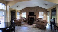 This single-floor space makes a strong impression with its thoughtfully designed living area, where a soaring vaulted ceiling adds a sense of openness and grandeur. The focal point of the room is a full-height exposed brick accent wall, which houses a classic fireplace topped with a mounted flat-screen television, creating a cozy yet functional centerpiece. The warm tones of the brick are complemented by golden-yellow walls, contributing to an inviting and relaxed atmosphere throughout. The living area is generously sized, comfortably accommodating a full suite of plush, earth-toned upholstered furniture, including sofas and armchairs arranged around a dark wood coffee table atop a decorative area rug. Large windows flanking the brick wall are fitted with woven wood blinds, allowing soft natural light to filter in while offering views of the surrounding greenery outdoors. The flooring consists of large-format stone-look tiles that add a grounded, durable quality to the space. Floor lamps provide warm ambient lighting, enhancing the room's cozy character. Overall, this space combines rustic charm with comfortable, practical design, making it well-suited for relaxed gatherings and everyday living.