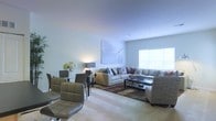 This single-floor residence offers an inviting open-concept layout that effortlessly blends the living and dining areas into one cohesive, functional space. The living area is anchored by a generously sized sectional sofa in neutral tones, complemented by a sleek glass-top coffee table and a patterned area rug that adds warmth and texture to the carpeted floor. A large window floods the room with natural light, enhancing the bright, airy atmosphere and highlighting the clean, smooth ceiling above. A piece of modern artwork adorns the wall, adding a touch of contemporary style to the space. Adjacent to the living area, the dining space features a glass-top dining table surrounded by modern upholstered chairs, along with a floral centerpiece that introduces a fresh, elegant accent. Ambient table lamps provide soft, warm lighting throughout. The overall design aesthetic is modern and understated, with a neutral color palette that creates a calm, welcoming environment. The seamless flow between zones makes this space well-suited for both everyday living and entertaining.