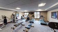 This fitness center offers a comprehensive workout environment designed to accommodate a wide range of exercise needs. The space is anchored by a cardio zone featuring multiple Matrix-brand treadmills and an elliptical trainer, along with a recumbent bike, all positioned near large windows that allow natural light to flow in and provide views of the surrounding outdoor area. A wall-mounted flat-screen TV is positioned for easy viewing during cardio sessions.

The strength training area is equally well-appointed, housing a multi-function cable machine and an additional strength station, providing options for a full upper and lower body workout. A dedicated free weights section includes a tiered dumbbell rack stocked with a range of weights, accompanied by adjustable workout benches ideal for a variety of exercises.

The room features a drop tile ceiling with recessed panel lighting, carpeted flooring with a geometric pattern, and a combination of neutral and bold navy blue accent walls that give the space a modern, polished feel. Brick accent walls add a textural contrast near the cardio equipment. The overall layout is open and spacious, allowing for comfortable movement between stations. Framed artwork on the walls contributes to an inviting, well-maintained atmosphere throughout the facility.