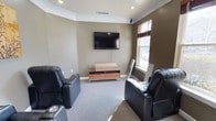 This single-floor space is designed as a dedicated media and entertainment room, offering a comfortable and functional layout for screen-based leisure. The room features warm, earthy taupe-toned walls accented by crisp white crown molding, which adds a touch of classic elegance to the overall aesthetic. Recessed ceiling lighting provides even, adjustable illumination throughout the space, complementing the natural light that filters in through a window dressed with horizontal blinds.

The focal point of the room is a wall-mounted flat-screen television, positioned at an optimal viewing height and paired with a mid-century modern-style wooden media console below, offering storage and display space. Two black leather recliner chairs with built-in cup holders are positioned facing the screen, providing dedicated, comfortable seating for an immersive viewing experience.

Soft, low-pile carpet in a neutral grey tone covers the floor, contributing to the room's quiet, relaxed atmosphere while also enhancing acoustics. The overall design balances practicality with comfort, making this space well-suited for home entertainment, movie viewing, or casual lounging.