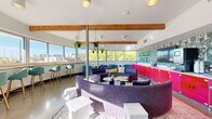 Vito's Lounge in Denver, Colorado is a stylishly designed communal space that masterfully blends bold aesthetics with an inviting, social atmosphere. The open-plan layout is defined by exposed wooden ceiling beams that add warmth and character against the crisp white walls and polished concrete floors. Wraparound floor-to-ceiling windows flood the interior with abundant natural light and offer sweeping panoramic views of the Denver skyline and surrounding treetops, creating a bright and airy environment throughout the day. At the heart of the lounge sits a distinctive curved, sectional sofa in rich purple upholstery, complemented by coordinating accent seating and low coffee tables that encourage conversation and gathering. Along the windowed perimeter, a built-in wooden ledge bar is lined with teal upholstered barstools, providing an ideal vantage point for enjoying the cityscape. A vibrant magenta cabinet unit anchors one wall beneath an eye-catching illustrated wallcovering and a mounted display screen, adding a playful, energetic focal point to the space. Hanging planters suspended from the beams introduce touches of greenery, softening the dynamic color palette. Teal armchairs and varied seating clusters throughout allow for both intimate and group settings, making Vito's Lounge a versatile and visually striking destination.