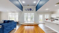 Located at Hamiltons Harbor in Lake Wylie, SC, this well-appointed single-floor space offers a seamless open-concept layout encompassing a living room, dining area, and fully equipped kitchen. The interior is defined by a striking navy blue and crisp white color palette, carried consistently throughout the space and complemented by warm wood-look flooring underfoot.

The entryway makes an immediate impression with soaring ceilings, bold navy accent walls, white crown molding, and a gold chandelier that adds an elegant touch. Large glass-panel entry doors flood the foyer with natural light and provide views of the surrounding landscape.

The kitchen is bright and modern, featuring white shaker cabinetry, a decorative tile backsplash, quartz countertops, a generous island, and a full suite of stainless steel appliances. Recessed lighting and crown molding maintain a clean, polished aesthetic throughout.

The living room is generously proportioned, with multiple large windows offering ample natural light and outdoor views. Deep navy upholstered seating anchors the space, creating a cohesive and inviting atmosphere.

The dining nook is intimate yet functional, featuring a built-in banquette with wainscoting beneath navy walls adorned with gold-framed artwork and a bright window that enhances the sense of openness. Together, these areas create a sophisticated, community-oriented environment.