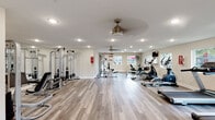 Located at 25650 Interstate 45 in Spring, TX, this single-floor fitness center is a comprehensive and thoughtfully designed workout facility. The space features durable hardwood-style flooring throughout, creating a clean and polished aesthetic that complements the bright, neutral-toned walls.

The strength training area is anchored by a multi-station cable machine and a variety of weight benches, accommodating a range of resistance and muscle-building exercises. Large wall-mounted mirrors reflect the space and allow users to monitor their form during workouts. A fire safety station is conveniently positioned nearby.

The cardio zone is generously equipped with multiple treadmills, elliptical trainers, stationary bikes — including a recumbent model — and a stair climber, providing diverse options for aerobic exercise. A wall-mounted television overlooks the cardio section, offering entertainment during workouts. A large industrial fan supports air circulation alongside ceiling fans fitted with integrated lighting.

Natural light filters in through several windows that frame views of the surrounding outdoor pool area, adding an open and airy feel to the space. Recessed lighting ensures the room is well-illuminated throughout. A small built-in counter and storage area near the entrance adds a practical touch to this fully functional, community fitness facility.