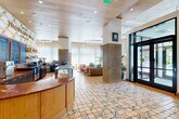 This generously sized single-floor hospitality space seamlessly blends a hotel lobby with a fully integrated café, Thousand Hills Coffee & Social, creating a vibrant and welcoming environment. The standout design element is the expansive, eclectic patterned tile floor that spans the entire space, featuring a rich mix of geometric and organic motifs in warm earth tones. Warm wood-paneled ceilings add an organic, grounding quality, complemented by recessed lighting and an elegant chandelier near the reception area. Exposed structural elements on one side of the ceiling offer an industrial contrast to the refined finishes elsewhere. Brick columns punctuate the open floor plan, adding architectural texture and character. The café counter, crafted from warm wood with a curved profile, anchors one side of the space alongside a glass display case and full barista station. A reception desk with a distinctive fringed base and an oversized art piece serves as a focal point, surrounded by lush indoor plants. Lounge seating areas feature leather sofas, upholstered chairs, and intimate built-in banquette nooks. Floor-to-ceiling sheer drapery diffuses abundant natural light from large windows and glass entry doors, while a curated gallery wall of framed artwork adds a personal, editorial touch throughout.