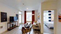 This well-appointed 2-bedroom, 2-bathroom residence is designed with modern living in mind, offering a thoughtful single-floor layout that maximizes comfort and functionality. The open living area serves as the heart of the home, featuring smooth hardwood-style flooring, crisp white walls, and large windows dressed with bold accent curtains that flood the space with natural light while adding a warm pop of color. A welcoming seating arrangement anchors the living room, which flows seamlessly toward the bedrooms. The bedroom visible from the main living area continues the clean, light aesthetic with ample space and additional window exposure. The layout provides a sense of openness and ease of movement throughout. Contemporary lighting fixtures, including a statement arc floor lamp, complement the modern design sensibility. The neutral palette throughout creates a versatile backdrop that feels both airy and inviting. With two full bathrooms and two generously sized bedrooms, this residence is ideal for those seeking both privacy and shared living space. The overall ambiance is bright, fresh, and thoughtfully designed, offering a comfortable and stylish place to call home.
