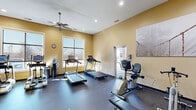 This fitness center at 3800 Enclave Ave in Sharonville, Ohio offers a well-appointed, single-floor exercise facility designed to support a comprehensive workout routine. The space features warm yellow and amber-toned walls complemented by a dark rubber flooring, creating an energetic yet functional atmosphere. Recessed lighting and a ceiling fan ensure the room remains well-lit and comfortable during workouts.

Natural light filters in through multiple windows, providing views of the surrounding exterior. The cardio area is equipped with several treadmills and stationary bikes, including both upright and recumbent models. A dedicated strength training section features a multi-function cable machine, a full dumbbell rack with a variety of weights, an adjustable workout bench, and an assortment of exercise balls. A wall-mounted mirror expands the visual sense of the space and supports form-checking during exercise.

Additional amenities include a wall-mounted television, a water cooler, and built-in lockers for secure storage of personal belongings. A wall-mounted shelf with sanitizing supplies promotes cleanliness. Keypad-secured entry doors provide controlled access. Motivational wall art adds a finishing touch to the overall design, making this fitness center a practical and inviting space for residents.