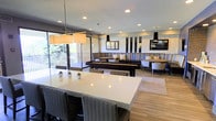 This single-floor communal space showcases a well-designed open-plan layout that seamlessly blends dining and lounge areas. At the forefront, a large white rectangular dining table surrounded by woven rattan chairs serves as a central gathering point, accented by small potted succulents and illuminated by a rectangular pendant light overhead. The light wood flooring and neutral-toned area rug contribute to the space's clean, contemporary aesthetic.

Moving deeper into the room, built-in banquette seating with bold striped blue and gold upholstery lines the walls, paired with dark wood tables and additional pendant lighting. A mix of upholstered chairs and booth-style seating creates distinct zones for both casual and formal gatherings. Recessed lighting throughout ensures consistent, warm illumination across the entire space.

Large sliding glass doors along one wall flood the interior with natural light and provide access to an outdoor balcony area with views of surrounding greenery. Decorative mirrors on the walls add depth, while the overall design strikes a balance between modern sophistication and comfortable, functional usability.