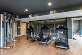 This single-level fitness center offers a comprehensive workout environment designed with both functionality and aesthetic appeal in mind. The space is divided into distinct workout zones, each thoughtfully outfitted to support a full range of exercise routines. The cardio area features multiple Cybex treadmills, an elliptical trainer, and a stationary bike, all arranged to allow ample room for movement. Adjacent to the cardio zone, a dedicated strength training area houses a multi-station cable machine alongside a free weight section equipped with a tiered dumbbell rack, adjustable bench, and additional accessories. Floor-to-ceiling mirrors line portions of the walls, enhancing the sense of space and supporting proper form during workouts. The interior is finished with warm hardwood-style flooring throughout, contrasted by deep charcoal and dark navy accent walls that lend a modern, polished atmosphere. Recessed lighting illuminates the space evenly, complemented by natural light streaming through large windows and glass panels. A decorative laser-cut metal screen partition adds an architectural design element, providing visual separation while maintaining an open feel. A wall-mounted flat-screen television is positioned for visibility during cardio sessions. Sliding glass doors open to an outdoor balcony, offering fresh air and exterior views. A small shelving unit provides storage for accessories and resistance bands.