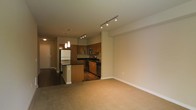 This single-floor space offers a thoughtfully designed open-concept layout that seamlessly connects the kitchen and living areas. The kitchen is a standout feature, outfitted with rich wood cabinetry, dark granite countertops, and a full suite of stainless steel appliances, including a refrigerator, stove, and built-in microwave. A central kitchen island provides additional prep space and serves as a natural dividing element between the cooking and living zones. Pendant lighting hangs above the island, adding a warm, focused glow to the workspace, while a track lighting system installed on the ceiling extends illumination throughout the open area. The living space is finished with plush wall-to-wall carpeting, creating a comfortable and inviting atmosphere. Neutral-toned walls and consistent finishes throughout contribute to a clean, cohesive aesthetic. A hallway visible near the entrance suggests additional private rooms beyond the main open area. The overall design balances functionality with a modern sensibility, making efficient use of the available space while maintaining an airy, open feel.