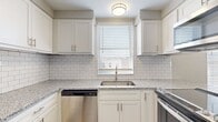 This two-story space is thoughtfully organized across two floors, with the ground level housing the combined kitchen and living area, a hallway, and an entry with a closet. The upper floor contains a bathroom and additional living space, connected via two staircases.

The ground floor kitchen is finished with crisp white shaker cabinetry extending to the ceiling, granite-speckled countertops, a classic subway tile backsplash, and stainless steel appliances including a dishwasher and range. A window above the sink introduces natural light into the cooking area. A granite-topped breakfast bar with a pass-through opening connects the kitchen to the living area, which features light wood-tone flooring and multiple windows fitted with blinds that fill the space with natural light. Neutral gray walls and clean white trim create a cohesive, modern palette throughout.

The upper floor features a striking vaulted ceiling that adds architectural character and a sense of openness to the space. The bathroom on this floor includes a tub-shower combination with sliding glass doors, white cabinetry, granite countertops, and matching wood-tone flooring, continuing the consistent design language found throughout the home.