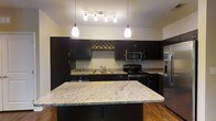 Located at 3245 Glen McConnell Parkway in Charleston, South Carolina, this single-floor residence offers a well-designed living space with thoughtful finishes throughout. The kitchen serves as a central highlight, showcasing rich dark cabinetry that contrasts elegantly with light granite countertops. A generously sized kitchen island with matching granite provides additional prep space and a natural gathering point. Stainless steel appliances, including a built-in microwave, electric range, and refrigerator, lend a modern, polished look to the space. A decorative wine rack built into the upper cabinetry adds a stylish and functional touch. Pendant lighting hangs above the island, complemented by a track lighting fixture that ensures the kitchen is well-lit for both cooking and entertaining. Warm hardwood flooring runs throughout, adding depth and warmth to the overall aesthetic. The layout is open and practical, allowing for seamless flow between the kitchen and adjoining living areas. The combination of dark cabinetry, granite surfaces, and stainless steel appliances creates a cohesive, contemporary design that is both functional and visually appealing, making this Charleston residence a refined and comfortable place to call home.