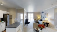 This single-floor residence offers a thoughtfully designed open-concept layout that seamlessly connects the kitchen, dining, and living areas into one cohesive, functional space. The interior is finished with neutral warm-toned walls and smooth ceilings accented by classic crown molding, creating a clean and refined atmosphere throughout.

The kitchen features dark granite-style countertops and flows directly into a bright dining area, where a round glass table provides an elegant spot for meals. The living room is generously sized, carpeted for comfort, and anchored by a light-toned sofa paired with a striking modern egg chair, offering a blend of contemporary and classic furnishings. A large sliding window at the far end of the living area fills the space with abundant natural light and provides views to the exterior, enhancing the open and airy feel of the room.

A doorway visible from the main living area leads to a private bedroom, which also benefits from natural light. The overall design palette — soft grays, warm whites, and coordinated accent pieces — lends the space a cohesive, move-in-ready aesthetic. The combination of practical layout, natural light, and stylish finishes makes this a comfortable and visually appealing single-level residence.