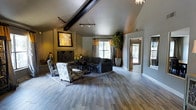 Located at Stonemill Village, this single-story space makes a strong first impression with its dramatic vaulted ceiling accented by an exposed dark wood beam, lending architectural character and a sense of openness to the main living area. The ceiling's angular pitch draws the eye upward and contributes to the spacious, airy feel of the room.

The flooring throughout is a contemporary wood-look material in a cool grey tone, providing a sleek, modern foundation that ties the space together cohesively. A fireplace with a decorative arched iron screen serves as a natural focal point along one wall, complemented by bold artwork displayed above it.

Track lighting runs along the vaulted ceiling, offering adjustable illumination across the room, while a statement chandelier is visible toward the entryway area. The living space is generously proportioned, accommodating a full seating arrangement including a sectional sofa and patterned accent chairs centered around a coffee table atop a layered area rug.

Large windows dressed with layered curtains invite natural light and frame views of the outdoors. A tall potted plant adds a lively natural element, and a secondary doorway with sidelight windows enhances connectivity to adjacent areas, reinforcing the open and welcoming ambiance of the space.