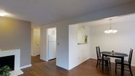This single-floor space offers a practical and well-organized layout designed to maximize functionality and flow. The interior is finished with rich, dark hardwood-style flooring that runs throughout, providing a warm contrast against the crisp white walls and neutral gray ceilings. The kitchen is partially enclosed, featuring a pass-through window that opens to the dining area, creating a sense of connectivity between spaces while maintaining a defined cooking zone. A full-size refrigerator anchors the kitchen, which benefits from ample wall space for additional appliances and cabinetry. The dining area is conveniently positioned adjacent to the kitchen pass-through, making it ideal for everyday meals and casual entertaining. A pendant light fixture overhead adds a subtle design touch to the dining space. The hallway connects the main living areas to additional rooms, offering clear sightlines and easy navigation throughout the unit. Textured ceilings add a classic finishing detail, while the consistent flooring and color palette throughout the space create a cohesive and tidy aesthetic. The overall ambiance is clean, understated, and move-in ready, offering a neutral backdrop well-suited to a variety of interior styles.