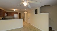 This two-story residence offers a practical and well-organized layout designed for comfortable everyday living. The main level showcases an open-concept design where the living area flows seamlessly into the kitchen, creating a spacious and connected environment. Light beige tile flooring extends throughout the main level, providing a clean, cohesive look that is both durable and easy to maintain.

The kitchen is equipped with stainless steel appliances, including a full-size refrigerator, and features warm wood-toned cabinetry complemented by a granite-topped breakfast bar that serves as both a prep space and a casual dining area. A decorative pendant light hangs above the bar, adding a subtle design touch. The living area is fitted with a ceiling fan with a light fixture, contributing to both air circulation and ambient lighting.

The main level also includes interior doors leading to additional rooms or utility spaces, with a staircase visible from the living area providing access to the second floor. A clerestory-style window on the upper portion of the interior wall allows light to filter between levels, enhancing the sense of openness. The overall ambiance is bright, neutral, and functional, making it a versatile space suited to a variety of living arrangements.