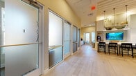 Located at 2950 Landover Blvd in Spring Hill, Florida, this single-floor professional office suite offers a thoughtfully designed workspace that blends functionality with coastal-inspired aesthetics. The layout includes six private offices accessible via two hallways, each enclosed by frosted glass doors with sleek aluminum frames that allow light to filter through while maintaining privacy.

The communal hub of the space centers around a striking white shaker-style bar island topped with a bright white surface and lined with dark-framed counter-height stools. Pendant lights hang above, complementing the built-in shelving unit that houses a mounted flat-screen display, a commercial coffee station, and decorative greenery. Light hardwood flooring runs throughout, enhancing the open and airy feel.

A welcoming lounge area features woven rattan furniture, a round ottoman, and an artful wall arrangement of woven basket décor, adding warmth and texture. A dedicated counter workspace with white cabinetry and marble-style backsplash provides additional utility. Vaulted and high ceilings with recessed lighting elevate the sense of space, while the warm neutral color palette and curated décor create a calm, professional ambiance well-suited for client-facing operations.