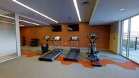 This fitness center offers a well-appointed workout space designed for cardio and general exercise. The room features bold burnt-orange accent walls that create an energetic atmosphere, complemented by neutral grey carpeting interspersed with orange geometric flooring sections that define the equipment zone. Overhead, sleek recessed linear LED lighting runs across the ceiling, providing bright, even illumination throughout the space. The cardio area is equipped with multiple treadmills, a stationary bike, and an elliptical trainer, all arranged in a row for easy access. Two wall-mounted flat-screen televisions are positioned at eye level on the orange feature wall, allowing users to stay entertained during workouts. A dumbbell rack with free weights is visible along the side wall, adding a strength-training component to the space. The clean, modern interior design is enhanced by white trim and a well-maintained layout that maximizes the available floor space. The overall ambiance is motivating and functional, with a contemporary aesthetic that balances bold color choices with practical fitness amenities.