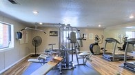 This fitness room offers a functional and well-organized workout space designed to support a variety of exercise routines. The room is anchored by a large multi-station cable and weight machine at its center, providing users with options for strength training across multiple muscle groups. A flat workout bench is positioned nearby, complementing the weight station for free-weight and resistance exercises. To one side, a stair climber and elliptical machine offer dedicated cardio training options. A standing floor fan is positioned along the wall, ensuring airflow and comfort during workouts. The flooring consists of light hardwood-style planks, with a protective mat laid beneath the weight equipment. Recessed lighting illuminates the space from a textured ceiling above, while a window allows a measure of natural light to filter in. Motivational posters and signage adorn the neutral-toned walls, reinforcing an encouraging atmosphere. The overall layout is practical and purposeful, making efficient use of the available space to accommodate both strength and cardiovascular training within a single, cohesive fitness environment.