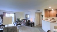 This single-floor residence offers a well-designed layout that balances comfort and functionality. The living area serves as the heart of the space, showcasing smooth, high ceilings with a centrally placed HVAC vent for year-round climate control. Soft, neutral wall tones create a clean and airy atmosphere throughout the room, complemented by plush wall-to-wall carpeting underfoot.

Natural light streams in through a large window dressed with elegant draping curtains, brightening the living space and creating a warm, inviting ambiance. The room provides ample space for a full seating arrangement, with clear sightlines that enhance the sense of openness. Decorative touches such as framed artwork and indoor plants add character and warmth to the environment.

A wide doorway leads from the living area into an adjacent hallway, connecting to additional rooms within the residence. A partial view of what appears to be a countertop or bar area is visible to the right, hinting at a nearby kitchen or dining space. The overall design is clean and straightforward, featuring practical finishes and a cohesive neutral palette that provides a versatile canvas for a variety of interior styles.