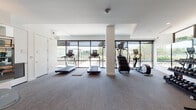 This contemporary fitness center is designed to support a comprehensive workout experience across a single, thoughtfully organized floor. The space is divided into distinct training zones, each outfitted with professional-grade equipment to accommodate a wide range of exercise routines.

The cardio zone features multiple treadmills, a stair climber, and an elliptical machine, all positioned in front of expansive floor-to-ceiling windows framed in dark metal. These large-pane windows flood the area with natural light and offer open views of the surrounding outdoor landscape, creating an energizing atmosphere for cardio workouts.

Adjacent to the cardio area, the strength training zone is equipped with multi-station cable machines, squat racks, weight benches, and a comprehensive free weights selection. Full-length mirrors line the walls, enhancing the sense of space while allowing users to monitor their form.

A separate cycling and studio area houses Echelon smart bikes with interactive screens, complemented by mirrored walls and a large flat-screen display. This dedicated space also includes storage lockers and a water fountain, adding functional convenience.

Throughout the facility, neutral-toned carpet flooring, recessed lighting, and a clean white palette contribute to a polished, modern aesthetic. The overall design balances openness with purpose, making it a well-appointed space for fitness and wellness.
