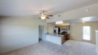 This single-floor space showcases an open-concept design that seamlessly connects the living area to the kitchen, creating a cohesive and functional layout. The living area is finished with plush wall-to-wall carpeting and features a ceiling fan with an integrated light fixture, contributing to both comfort and air circulation. Neutral wall tones throughout lend a clean, versatile backdrop suited to a variety of interior styles.

The kitchen is well-appointed with warm-toned wooden cabinetry offering ample storage. A full suite of stainless steel appliances is included, featuring a French door refrigerator, an electric range, and a built-in microwave. The kitchen island doubles as a breakfast bar with a built-in sink, providing additional prep space and casual dining potential. Overhead fluorescent lighting ensures the kitchen area remains bright and practical.

A hallway visible from the main living space leads to additional rooms, suggesting a thoughtful floor plan that maintains privacy while keeping common areas open. The space benefits from multiple ceiling vents, supporting effective climate control throughout. Overall, the unit offers a functional, move-in-ready environment with a straightforward, practical aesthetic.