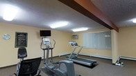 This single-floor fitness room offers a straightforward and functional space designed to accommodate cardio-focused workouts. The room is equipped with multiple treadmills and at least two elliptical cross-trainers, providing users with a variety of aerobic exercise options. A wall-mounted flat-screen television is positioned at an ideal viewing height, allowing users to stay entertained during workouts.

The space features a textured ceiling with dark decorative wooden beams that add a subtle architectural character to the room. Overhead fluorescent lighting provides adequate illumination throughout. The walls are painted in a warm, neutral yellow tone, contributing to an inviting atmosphere. A vertical blind-covered window along one wall allows for natural light while maintaining privacy.

A wall-mounted air conditioning unit ensures the room stays climate-controlled during use. The flooring is covered in low-pile carpet, which provides a degree of cushioning underfoot. Baseboard heating elements line the lower portions of the walls, suggesting the space is designed for year-round use. Overall, the room is compact yet efficiently arranged to maximize usable workout space.