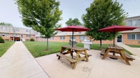 This single-floor property features a welcoming outdoor communal area designed for relaxation and social gatherings. The paved patio serves as the centerpiece of the outdoor space, accommodating multiple wooden picnic tables, each shaded by bold red market umbrellas that provide comfortable relief from the sun. Two stainless steel gas grills are conveniently positioned nearby, making the area well-suited for outdoor cooking and entertaining. The patio is bordered by a generous expanse of well-kept green lawn, which adds a sense of openness and tranquility to the setting. Mature deciduous trees are thoughtfully spaced throughout the grounds, offering natural shade and a pleasant, park-like atmosphere. The surrounding brick building with neutral siding provides a clean, residential backdrop that frames the outdoor area. Overall, the space strikes a balance between functional amenity and inviting outdoor living, offering residents a dedicated area to gather, dine, and enjoy the outdoors in a comfortable and well-appointed setting.