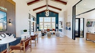 Located at 100 Hive Dr in Charlotte, NC, this single-floor community amenity center offers a thoughtfully designed space for residents to gather, stay active, and relax. The living room serves as the heart of the interior, boasting soaring vaulted ceilings with exposed wood beams, large windows that flood the space with natural light, and a striking floor-to-ceiling tiled fireplace. Bold navy accent walls and light hardwood flooring lend the room a warm yet modern character. Adjacent to the living area, the kitchen is bright and contemporary, featuring white cabinetry, open shelving, a large central island with seating, pendant lighting, and a vibrant Charlotte cityscape mural that adds a local flair. The dedicated exercise room is well-appointed with professional-grade strength equipment, free weights, and ceiling fans, all set on durable dark flooring with ample natural light from surrounding windows. Outside, the amenity grounds shine with a manicured artificial turf lawn strung with bistro lights, a covered patio furnished with lounge seating and ceiling fans, a fire pit area, and a fenced pickleball court surrounded by thoughtful landscaping. Together, the indoor and outdoor spaces create a dynamic and inviting community hub.