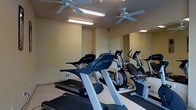 This fitness room is a well-appointed exercise space designed to meet a variety of cardio training needs. The room features a selection of True-brand cardio equipment, including treadmills and elliptical machines, arranged to allow comfortable use and easy movement between stations. A large wall mirror spans one side of the room, creating a sense of expanded space while allowing users to monitor their form during workouts. The ceiling is outfitted with two ceiling fans that provide consistent air circulation, helping to keep the environment comfortable during exercise sessions. Overhead fluorescent lighting ensures the room is well-lit throughout. The light-colored walls and neutral flooring contribute to a clean, focused atmosphere that is conducive to exercise. The layout is practical and efficient, making the most of the available space without feeling cramped. Overall, this fitness room offers a functional and comfortable environment for residents or users seeking a convenient on-site workout facility.