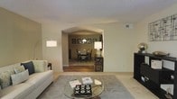 This single-story residence is designed with a practical and welcoming layout that promotes easy movement between living spaces. The living room serves as the central gathering area, featuring neutral-toned carpet, a textured ceiling, and warm beige walls that create a calm, understated ambiance. A glass-top coffee table anchors the seating area, complemented by a sleek arc floor lamp that adds both style and functional lighting.

A distinctive arched passageway connects the living room to the dining area, serving as an architectural focal point that adds character while maintaining an open feel. The dining room features hardwood flooring, a chandelier overhead, and a dark wood dining set surrounded by framed wall art, lending the space a polished, curated appearance. A console table with a table lamp in the transitional space adds warmth and continuity between the two rooms.

The consistent neutral color palette and coordinated furnishings throughout reinforce a cohesive interior aesthetic. Natural and ambient lighting work together to keep the space feeling bright and inviting. Overall, the home offers a well-connected, comfortable environment suited for both everyday living and entertaining.