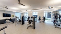This contemporary fitness center is designed to provide a comprehensive workout environment within a bright, open layout. The space is outfitted with a full suite of True-brand exercise equipment, including treadmills, elliptical trainers, stationary upright and recumbent bikes, and a selection of strength training machines — offering users a well-rounded range of exercise options. The light-toned flooring and crisp white ceilings contribute to a clean, modern aesthetic throughout. Large windows line the perimeter of the space, inviting abundant natural light and offering views of the outdoor surroundings, creating an open and energizing atmosphere. Multiple ceiling fans are suspended overhead, ensuring comfortable air circulation across the entire floor. Linear pendant lighting supplements the natural light, maintaining consistent brightness throughout the room. The single-level layout provides a spacious, uncluttered environment that allows for easy movement between equipment. The overall design balances functionality with a polished, well-maintained appearance, making it a welcoming facility for a variety of fitness routines and skill levels.