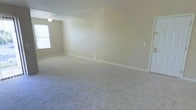 This single-floor residence in the United States presents a straightforward and functional layout designed for comfortable everyday living. The interior is finished with wall-to-wall plush carpeting in a soft, neutral tone that extends throughout the main living area, providing a warm and quiet underfoot experience. The walls are painted in a light beige hue, creating a bright and cohesive atmosphere that pairs well with a variety of furnishing styles. A window along one wall allows natural light to filter into the space, enhancing the open and airy feel of the room. The ceiling features a simple flush-mount light fixture, and a ceiling-mounted vent indicates a central HVAC system for year-round climate control. White baseboards and a light switch plate are neatly integrated into the walls, reflecting a clean and well-maintained finish throughout. The room offers generous open floor space, providing flexibility for a range of furniture arrangements to suit different lifestyle needs. A doorway visible at the edge of the space suggests additional rooms beyond, contributing to the overall flow of the home. The space is move-in ready, presenting a clean, neutral canvas that is easy to personalize.