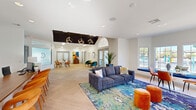 This single-story clubhouse facility, branded as Essena, offers a well-appointed community gathering space designed with a contemporary aesthetic throughout. The expansive main lounge serves as the heart of the space, featuring herringbone-patterned light wood flooring, soaring ceilings accented by a bold black ceiling panel with geometric cube pendant lighting, and an arched transom window that floods the room with natural light. Vibrant, colorful furnishings — including a navy sofa, jewel-toned accent chairs, and warm orange ottomans — are arranged over a vivid area rug, creating an inviting and energetic atmosphere. A dedicated reception area features a warm wood desk, textured diamond-relief wall tile, and frosted glass panels etched with the Essena branding, lending a polished, professional feel. The game and social room is bright and airy, with high ceilings, clerestory windows, a wall-mounted television, foosball table, and a built-in wet bar with wood cabinetry — ideal for social gatherings. The indoor pool and spa area features a tiled hot tub, marble-look tile wainscoting, a slatted ceiling detail, and access to private shower stalls, completing this well-rounded amenity space.