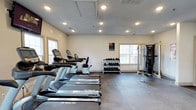This fitness center offers a thoughtfully designed workout environment built to support a full range of exercise routines. The space features durable, high-quality flooring suited for heavy equipment and high-traffic use, providing a stable and comfortable surface throughout.

A row of True-brand treadmills lines one side of the room, offering ample cardio options for users of all fitness levels. Alongside the treadmills, elliptical machines and an upright stationary bike provide additional low-impact cardiovascular options. On the strength training side, a well-organized free weight rack holds a complete set of dumbbells, neatly arranged for easy access. A multi-functional cable and weight machine anchors the strength training zone, enabling a wide variety of resistance exercises.

Natural light filters in through multiple windows positioned along the walls, creating a bright and energizing atmosphere. Recessed ceiling lighting supplements the natural light, ensuring the space remains well-illuminated at all hours. The walls are finished in a clean, neutral tone, contributing to an open and focused workout environment. Ventilation is supported by ceiling vents, keeping the space comfortable during intense exercise sessions. Overall, this fitness center is a practical and inviting space designed to meet diverse fitness needs.