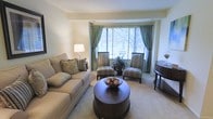 This single-floor residence offers a thoughtfully designed living space that balances comfort and elegance. The living room serves as the centerpiece of the home, featuring a plush, neutral-toned sofa accented with decorative pillows, a pair of classic striped armchairs, and a rich dark-wood round coffee table that anchors the seating arrangement. A decorative bowl centerpiece adds a refined finishing touch to the ensemble. Large bay-style windows dressed with sage green drapes and horizontal blinds allow an abundance of natural light to fill the room while offering a pleasant view of the surrounding greenery outdoors. The soft, wall-to-wall carpeting in a warm beige tone contributes to a cozy and inviting ambiance throughout. A stylish table lamp provides warm accent lighting, complementing the neutral walls and creating a relaxed atmosphere. An elegant side table near the window adds additional functionality and visual interest. The overall aesthetic of the space is calm and sophisticated, with a harmonious blend of earthy tones and traditional furnishings that make it feel both welcoming and polished.