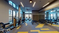 This single-floor fitness center is designed to accommodate a comprehensive workout experience within a generously proportioned, open layout. The space features soaring double-height ceilings that enhance the sense of openness, complemented by an abundance of natural light streaming through large, multi-pane windows lining the walls and clerestory windows positioned above, offering views of the surrounding exterior.

The flooring throughout consists of modular carpet tiles in a bold grey and yellow geometric pattern, adding a dynamic visual element to the space. Linear LED lighting runs along the ceiling, providing consistent, bright illumination throughout.

The fitness center is divided into functional zones. A dedicated cardio area houses treadmills, elliptical trainers, and a rowing machine positioned directly in front of the large windows. A strength training zone features a multi-station cable machine and additional resistance equipment. A free weights area includes a full dumbbell rack paired with adjustable benches and floor space for stretching or functional training.

Full-length mirrors along one wall amplify the sense of space. A dedicated entry area features dual water fountains, a decorative paneled accent wall with a geometric design, and posted fitness center guidelines, completing this well-appointed facility.