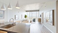 Located in the heart of Nashville at 805 Church Street, this modern one-bedroom, one-bathroom high-rise residence offers a refined urban living experience defined by clean lines, quality finishes, and breathtaking city views. The open-concept kitchen and living area serves as the heart of the home, where floor-to-ceiling windows flood the space with natural light and frame dramatic panoramic views of the Nashville skyline. The kitchen is appointed with stainless steel appliances, sleek cabinetry, a dark stone countertop island with bar seating, and pendant lighting overhead. Light hardwood flooring flows throughout, complementing the modern, neutral palette. Exposed concrete ceiling elements add an industrial character that contrasts beautifully with the bright, airy atmosphere. The bedroom continues the floor-to-ceiling window theme, offering sweeping views of the downtown skyline as a serene backdrop. Two closets and a dedicated laundry area provide practical storage solutions. The bathroom features a clean, contemporary design with a stone-topped vanity, an illuminated mirror, and large-format tile flooring. The welcoming entrance leads smoothly into the open living spaces, making the layout feel efficient and connected throughout.