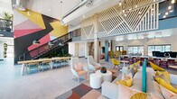 This expansive single-floor community clubhouse is a thoughtfully designed social hub that blends modern industrial architecture with bold, contemporary interiors. Soaring double-height ceilings with exposed ductwork and structural beams lend an open, loft-like character throughout, while floor-to-ceiling glass walls and oversized windows flood the space with abundant natural light and offer clear views to the surrounding outdoor landscape.

The main gathering area serves as the heart of the space, anchored by a dramatic large-scale geometric mural in vivid yellow, red, and black tones. A welcoming reception desk greets visitors upon entry beneath a striking "hello" accent wall. Multiple distinct seating zones are furnished with a vibrant mix of modular sofas, high-back accent chairs in mustard and teal, and colorful geometric-patterned area rugs, encouraging both casual lounging and social interaction.

A central kitchen and bar island provides a communal workspace and social counter, complemented by warm wood cabinetry, pendant lighting, and a beverage station. A mezzanine level overlooks the main floor, accessible by a statement staircase adorned with a bold mural. Tucked private meeting rooms with glass partitions offer quieter workspaces. Decorative wood-slat wall panels, a built-in shelving unit with a fireplace, and curated artwork complete the polished, energetic ambiance of this dynamic community space.