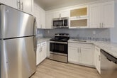 Located at 107 Wartburg Ave in Copiague, NY, this single-floor unit offers a thoughtfully designed open-concept layout that seamlessly connects the living area and kitchen. The kitchen is a standout feature, showcasing white shaker-style cabinetry with frosted glass upper panels, granite-look countertops, a classic subway tile backsplash, and a full suite of stainless steel appliances including a refrigerator, range, and over-the-range microwave. A peninsula with matching countertop provides additional prep space and defines the kitchen from the expansive living area. Light wood-tone luxury vinyl plank flooring runs throughout, lending warmth and cohesion to the entire space. Multiple windows invite natural light into both the living area and the bedrooms, offering views of the surrounding neighborhood. The bedrooms are finished in soft, neutral blue-gray tones and feature sliding closet doors with built-in rod storage. Baseboard heating is present throughout. A dedicated laundry closet houses a stacked washer and dryer unit for added convenience. Clean white trim, neutral walls, and contemporary finishes throughout give the space a fresh, move-in-ready character.