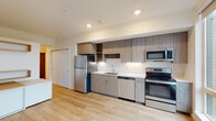This single-floor unit showcases a clean, contemporary design throughout. The kitchen is a standout feature, equipped with a full suite of stainless steel appliances including a refrigerator, electric range, built-in microwave, and dishwasher. Flat-front cabinetry in a warm wood-grain finish extends from floor to ceiling, complemented by open shelving for display or additional storage. Light-colored countertops provide ample workspace, and recessed lighting illuminates the area efficiently.

The open layout connects the kitchen seamlessly to the living space, both finished with light hardwood flooring that contributes to a bright, airy atmosphere. The living area benefits from a large floor-to-ceiling sliding window that floods the room with natural light and frames a view of the surrounding cityscape. A wall-mounted mini-split unit provides climate control.

The bathroom maintains the unit's modern aesthetic with a wood-toned vanity, white quartz-style countertop, undermount sink, and a full-width mirror. A walk-in shower with a rod and clean white surround completes the bathroom. Throughout the unit, the neutral color palette, clean lines, and consistent material choices create a cohesive and functional living environment.