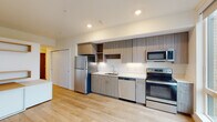 This single-floor unit showcases a clean, contemporary design throughout. The kitchen is a standout feature, equipped with a full suite of stainless steel appliances including a refrigerator, electric range, built-in microwave, and dishwasher. Flat-front cabinetry in a warm wood-grain finish extends from floor to ceiling, complemented by open shelving for display or additional storage. Light-colored countertops provide ample workspace, and recessed lighting illuminates the area efficiently.

The open layout connects the kitchen seamlessly to the living space, both finished with light hardwood flooring that contributes to a bright, airy atmosphere. The living area benefits from a large floor-to-ceiling sliding window that floods the room with natural light and frames a view of the surrounding cityscape. A wall-mounted mini-split unit provides climate control.

The bathroom maintains the unit's modern aesthetic with a wood-toned vanity, white quartz-style countertop, undermount sink, and a full-width mirror. A walk-in shower with a rod and clean white surround completes the bathroom. Throughout the unit, the neutral color palette, clean lines, and consistent material choices create a cohesive and functional living environment.