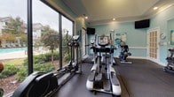 Located at 1341 Ellis Forest Rd in Kernersville, NC, this well-equipped fitness center offers a comprehensive workout environment within a single-floor layout. The space is finished with soft aqua-toned walls, crisp white crown molding, and dark rubber flooring designed to withstand heavy use. Elevated ceilings with recessed lighting and a tray ceiling detail add a sense of openness throughout.

The cardio area is lined with treadmills, ellipticals, and stationary bikes, positioned to take full advantage of the large floor-to-ceiling windows that frame views of the outdoor pool and surrounding landscaping. Wall-mounted televisions are conveniently placed for entertainment during workouts.

The strength training section features a multi-functional cable machine, a Smith machine with weight plates, a full dumbbell rack, and an assortment of medicine and stability balls for functional fitness. A large wall mirror spans one side of the room, enhancing the sense of space and allowing for form monitoring.

Natural light flows generously through multiple oversized windows and a glass-paneled exterior door, connecting the interior to the outdoor environment. Safety features including a fire extinguisher and clearly marked exit signage are present throughout. A water fountain is also accessible within the space, completing this thoughtfully appointed fitness facility.